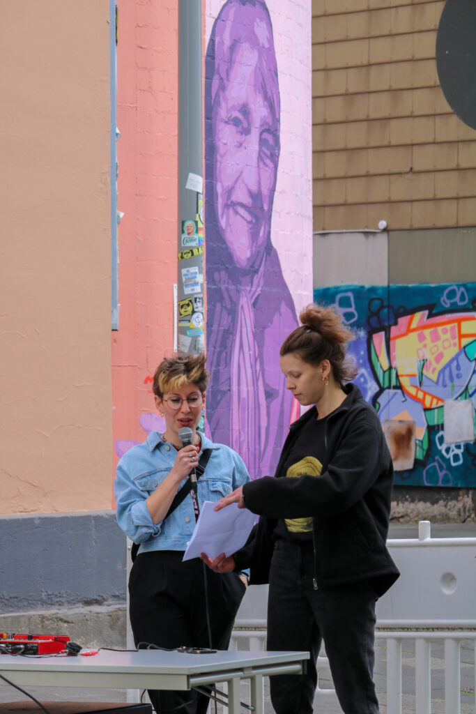 Zwei Personen halten eine Rede, im Hintergrund sieht man ein Wandgemälde